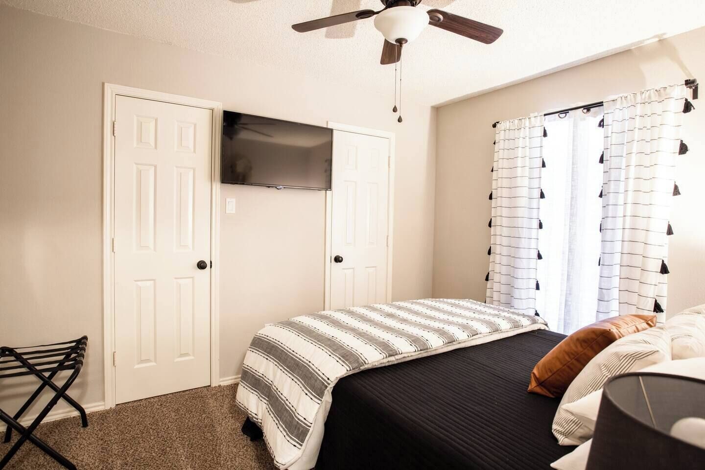 Quarto com cama, ventilador de teto, televisão fixada na parede e janela com cortinas.