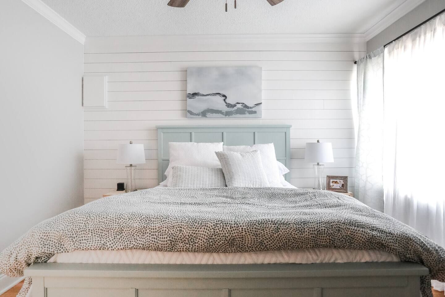 Una camera da letto con un letto, una testiera, due lampade da comodino, un quadro incorniciato e una finestra con tende.