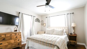 Ein Schlafzimmer mit Bett, Holzkommode, Nachttisch, Lampe und einem an der Wand montierten Fernseher.