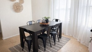 Zona pranzo con tavolo in legno scuro, sedie nere, lampadario nero e tenda bianca.