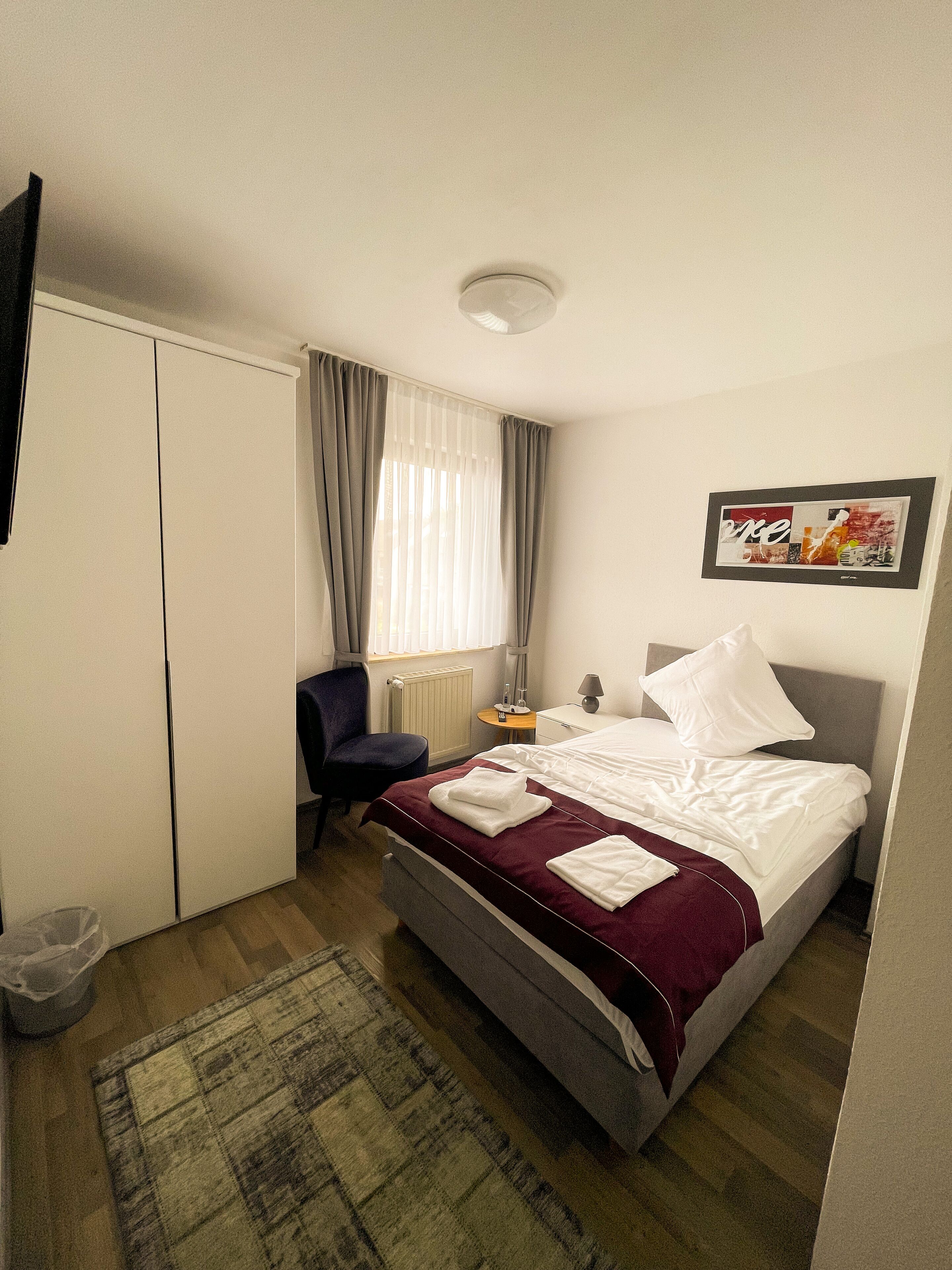 Un cuarto de hotel con cama, escritorio, silla, ventana con cortinas, radiador y un cuadro en la pared.