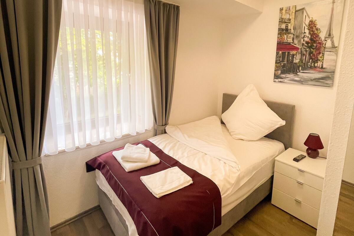 Hotellihuone, jossa on sänky, yöpöytä, lamppu, taulu ja ikkuna, jossa on verhot.