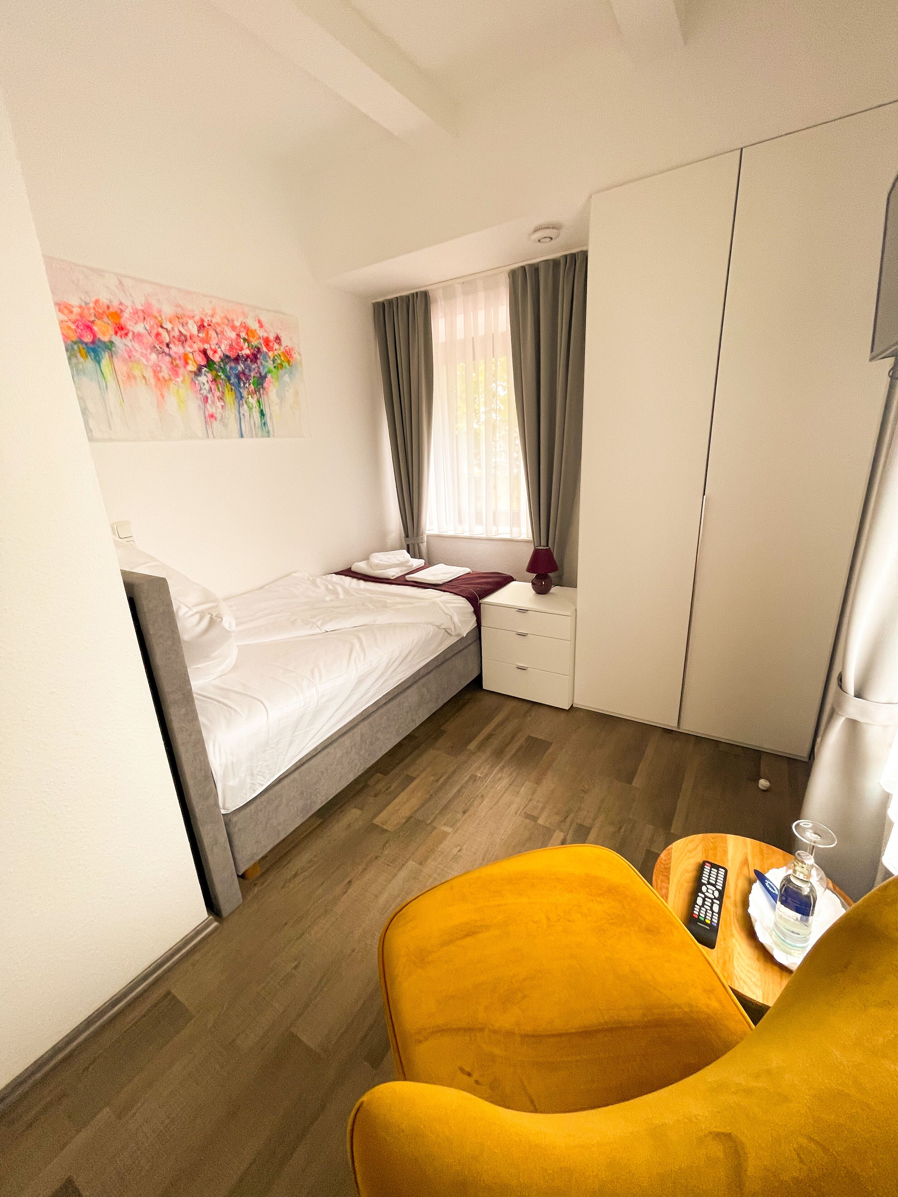 A small, well-lit room with a bed, a nightstand, a chair, a table with a remote control and a bottle, and a painting on the wall.
