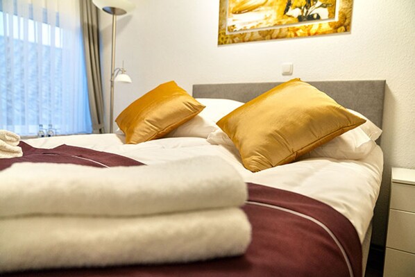 Una cama bien hecha con ropa de cama blanca y granate, dos almohadas doradas y una toalla blanca doblada.