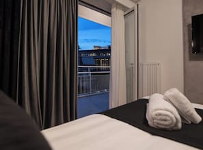A hotel room with a bed, a view of a building through an open door, and a balcony with a railing.
