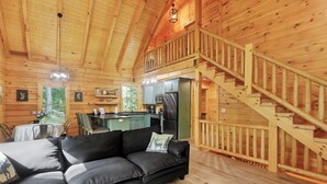 Een houten blokhut interieur met een zwarte leren bank, een eettafel met stoelen, een keuken en een houten trap.