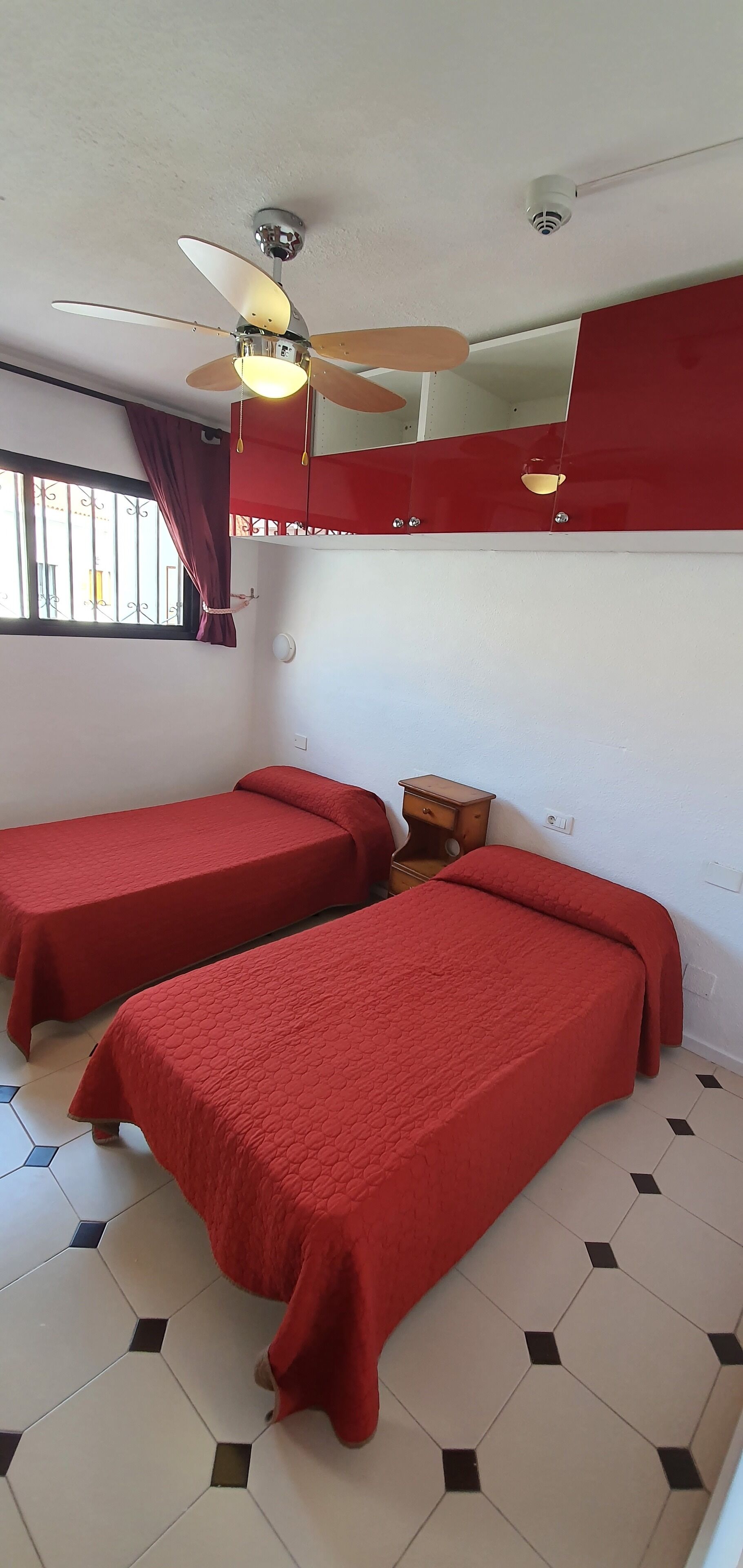 Una habitación con dos camas, un ventilador de techo, una ventana con cortinas y una pequeña mesa de madera.