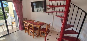 Uma área de refeições com mesa e cadeiras de madeira, uma televisão fixada na parede e uma escada espiral vermelha.