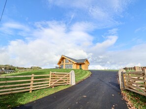 A wooden house with a sloped roof and large windows, surrounded by a grassy area and a paved road. - Pen Bryn (Ruthin)