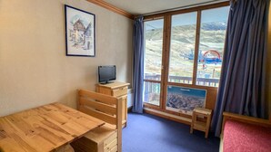 Et værelse med et træbord, et fjernsyn på et stativ, et vindue med udsigt til et bjerglandskab og et billede i ramme på væggen.