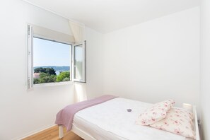 A bedroom with a bed, a window with a view of greenery and buildings, and a white wall.