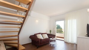 Una sala de estar con escalera de madera, un sofá marrón, una mesita blanca y un balcón con cortinas blancas.