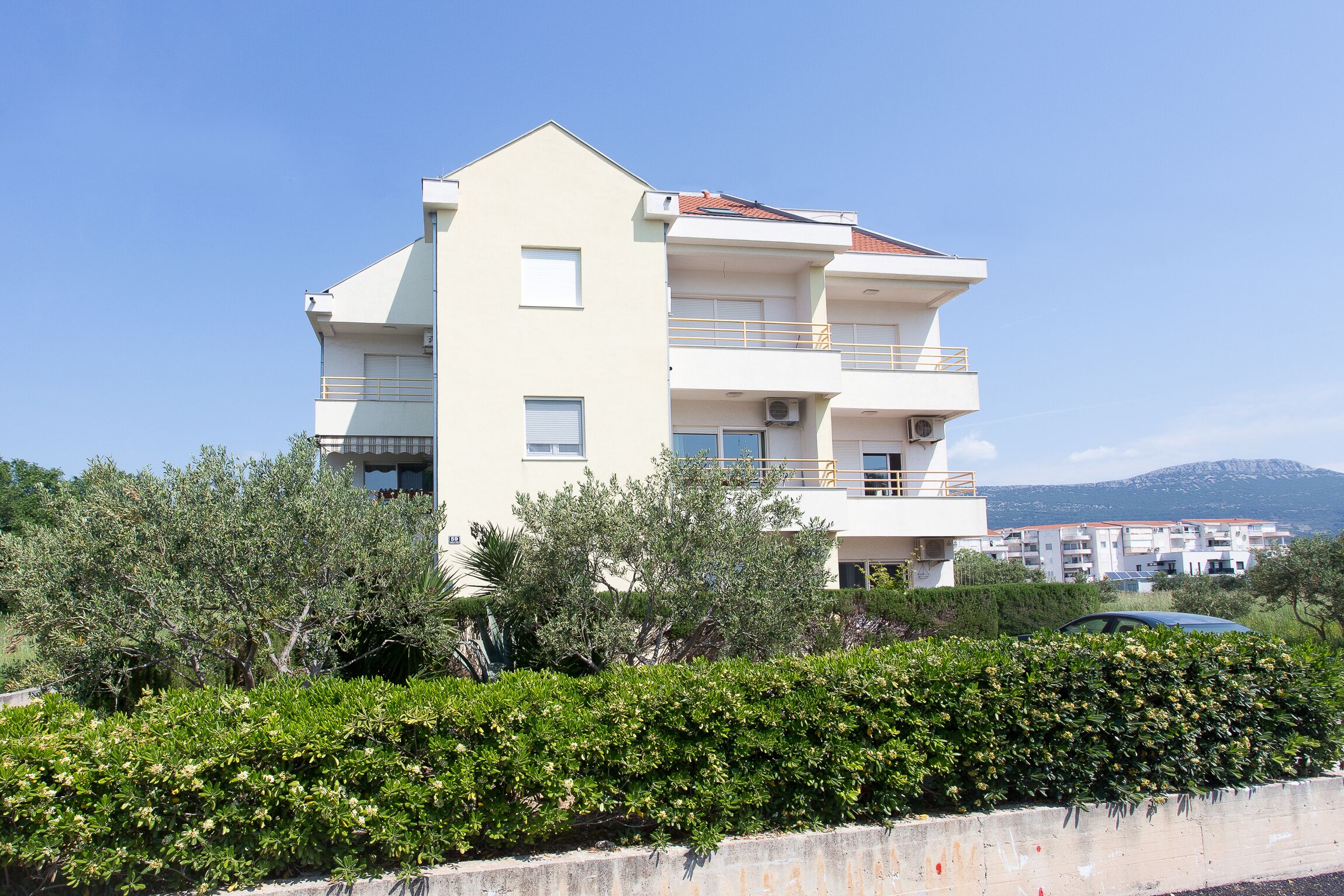 A multi-story residential building with balconies, surrounded by greenery and a clear sky.