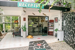 Een receptiegedeelte met een bordje met daarop 'RECEPTIO', een glazen toegangsdeur, een kleine zithoek met stoelen en een potplant.