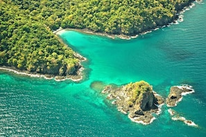 Luchtbeeld van een kustgebied met helder turquoise water, rotsformaties en weelderige groene begroeiing.