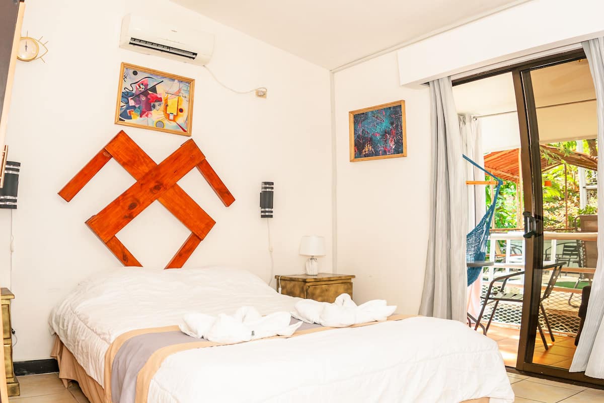A hotel room with a bed, a ceiling fan, an air conditioning unit, a small table with a lamp, and a view of the outdoors through a sliding glass door.
