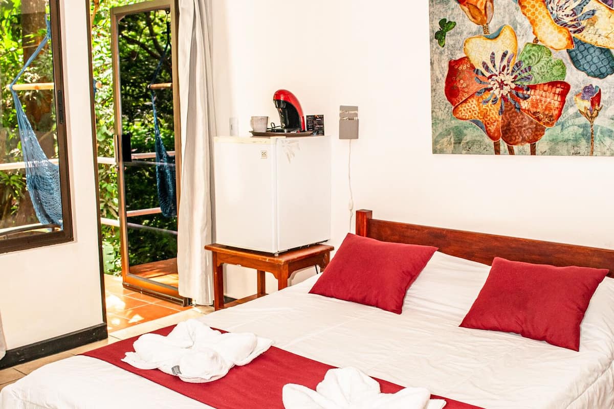 A hotel room with a bed, red and white bedding, a painting on the wall, and a view of greenery through the window.