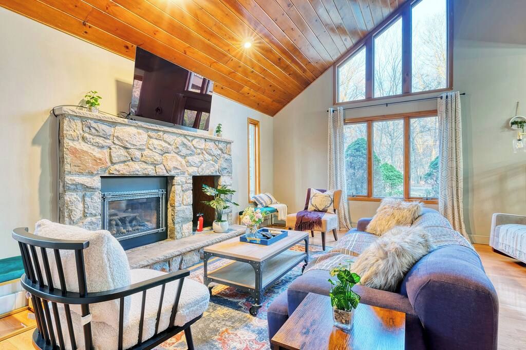 A cozy living room with a stone fireplace, wooden ceiling, and large windows.