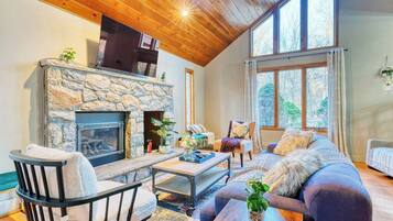 A cozy living room with a stone fireplace, wooden ceiling, and large windows.