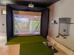 A projection screen displaying a golf course selection interface, a golf club rack, and a mat on the floor. - Antler Ridge: Hot Tub Golf Simulator Game Room (Penn Forest Township)