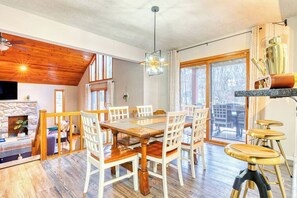 A dining area with a wooden table and white chairs, a stone fireplace, and a large window with a view. - Antler Ridge: 3BDR w/ Hot Tub & Game Room (Penn Forest Township)