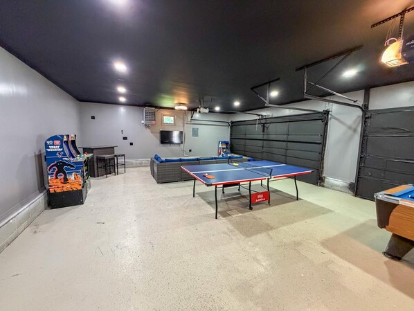 A room with a ping pong table, a sofa, a small table, and a gaming machine.