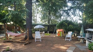 Ein Garten mit einer Hängematte, weißen Stühlen, einem kleinen Spielhäuschen und einem Grillplatz.