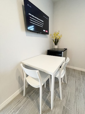 A modern dining area with a white table and chairs, a flat-screen TV mounted on the wall, and a small black side table with a vase of yellow flowers.