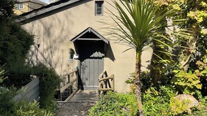 A building with a wooden staircase leading to a dark door, surrounded by greenery and a palm tree.
