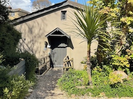A building with a wooden staircase leading to a dark door, surrounded by greenery and a palm tree.
