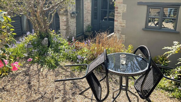 A patio with a glass table and metal chairs, surrounded by a garden with various plants and flowers.