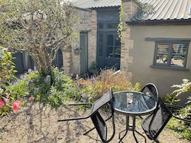 A patio with a glass table and metal chairs, surrounded by a garden with various plants and flowers.