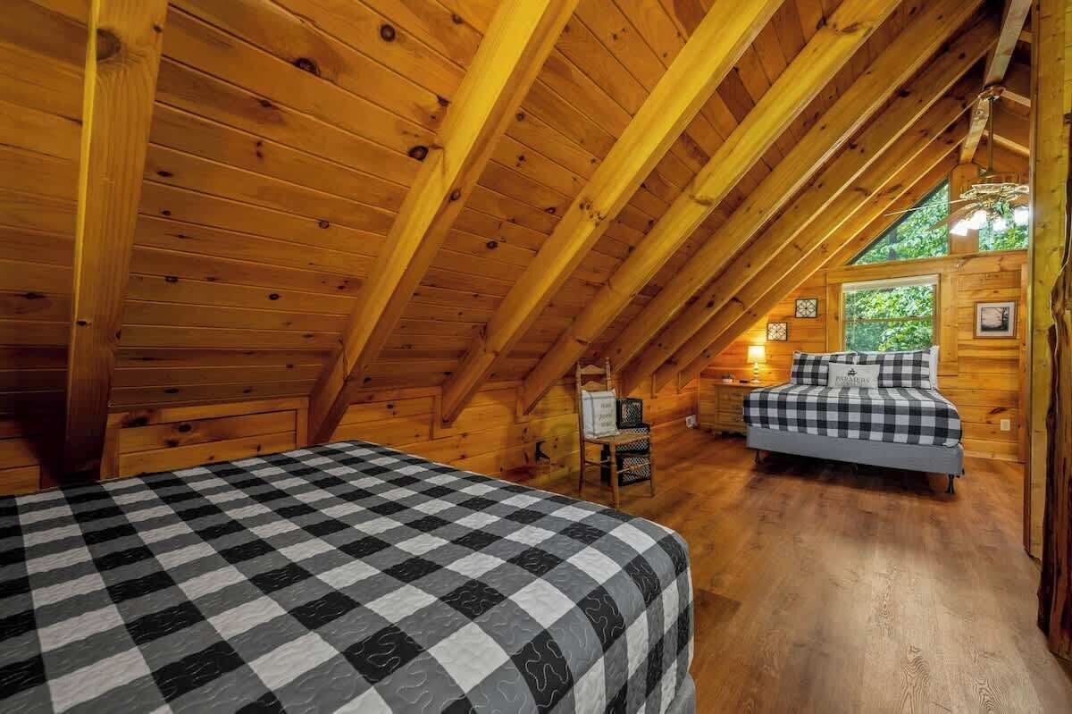 Une chambre mansardée chaleureuse avec des poutres apparentes, un lit à carreaux, une petite table et une fenêtre donnant sur un paysage verdoyant.