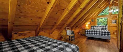 Une chambre mansardée chaleureuse avec des poutres apparentes, un lit à carreaux, une petite table et une fenêtre donnant sur un paysage verdoyant.