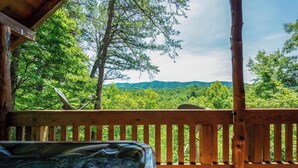 A wooden deck with a hot tub, surrounded by lush greenery and a view of distant hills. - Nice View! Log Cabin,Fireplace,Hot Tub,Pool Table (Sevierville)