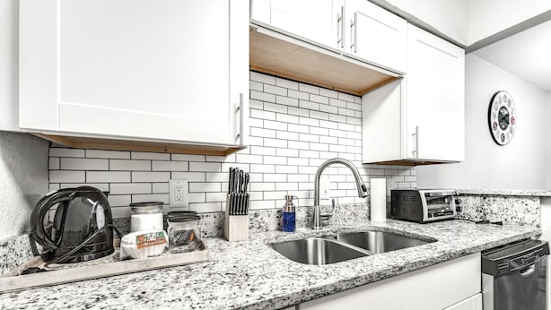 A modern kitchen with white cabinets, a granite countertop, a stainless steel sink, a toaster, and a clock on the wall.