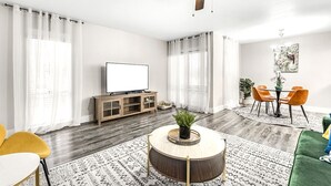 A modern living room with a round coffee table, a flat-screen TV, a ceiling fan, and a dining area with orange chairs.