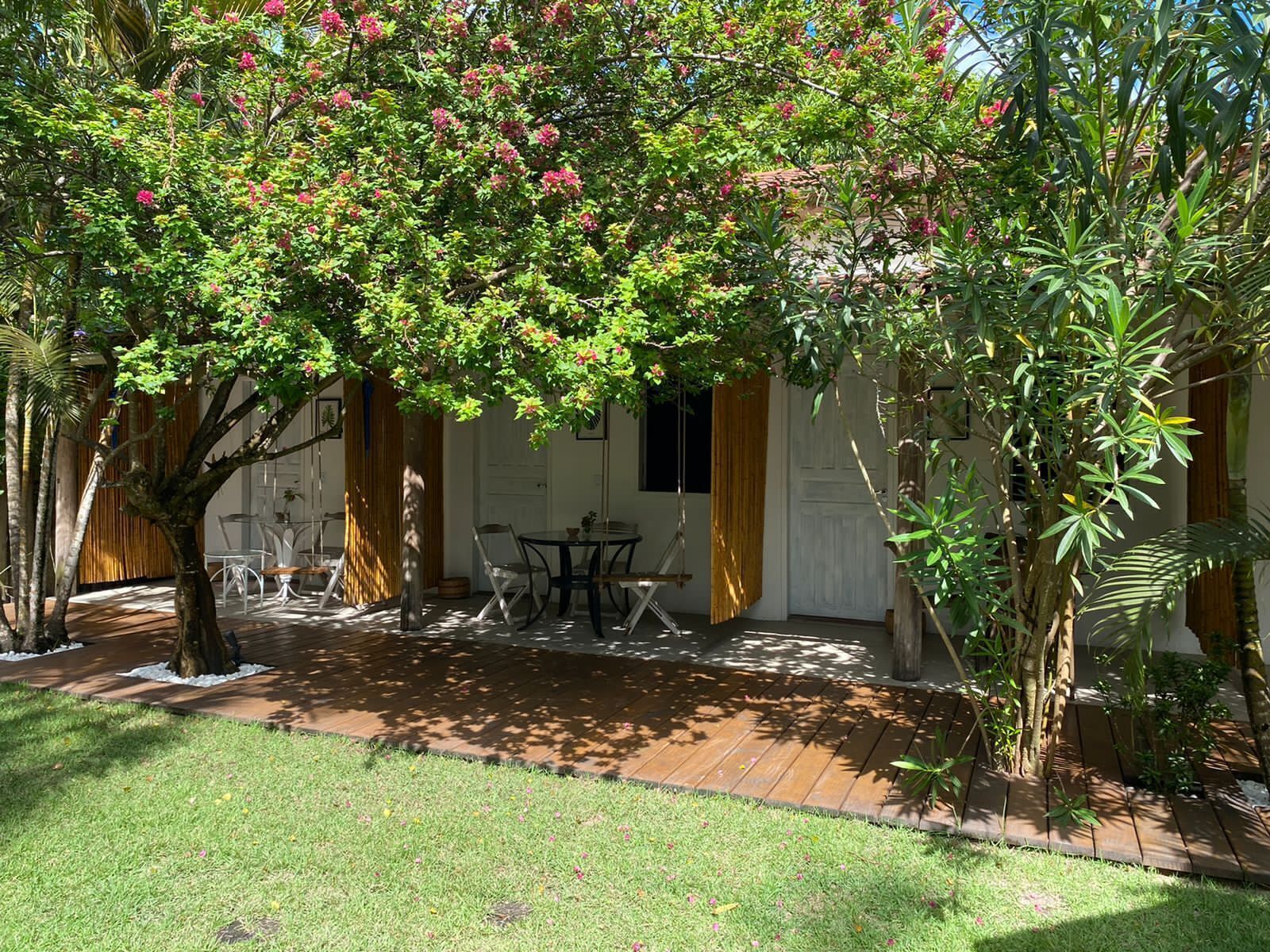 Un patio con mesa y sillas, rodeado de exuberante follaje y un árbol con flores rosadas.
