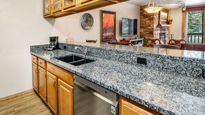 A kitchen with granite countertops, wooden cabinets, a dishwasher, and a stone fireplace. - Mountain Modern Beaver Creek Village Condo, Ski-In and Out, w/ Hot Tub and Views (Beaver Creek)