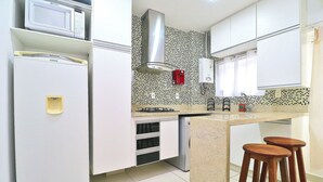 A kitchen with white cabinets, a microwave, a gas stove, and a dishwasher. There are two wooden stools at the counter. - Rio Spot Homes MC C055 (Rio de Janeiro)