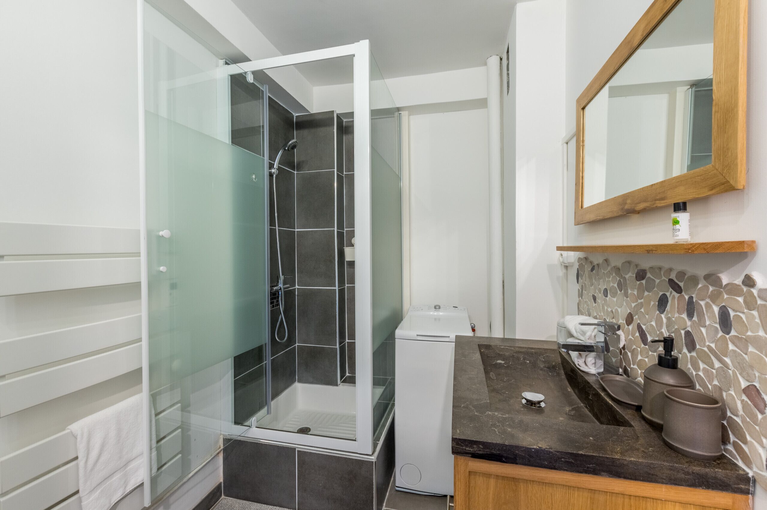 A modern bathroom with a glass-enclosed shower, a white washing machine, a granite countertop, and a stone mosaic backsplash.