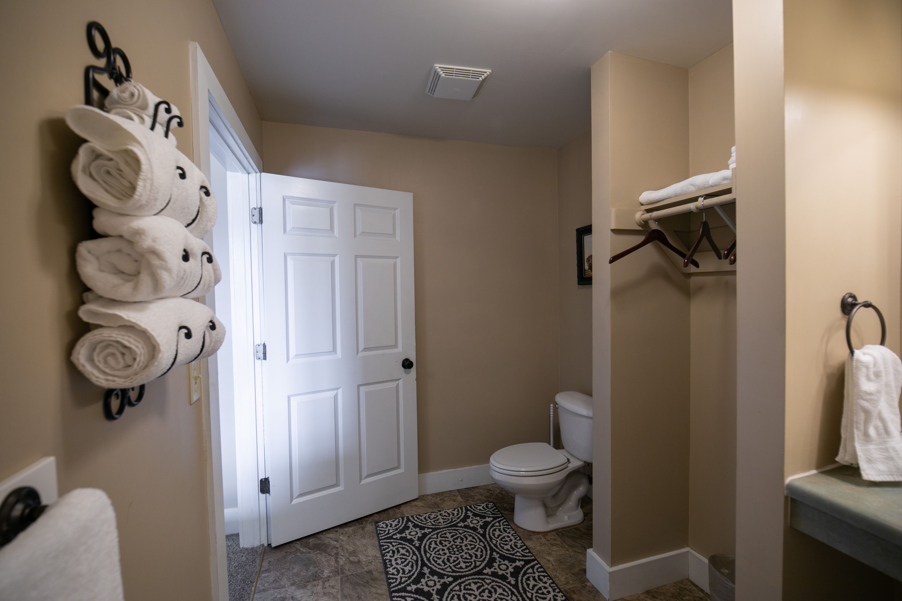 A bathroom with a toilet, a towel rack with rolled towels, and a patterned rug.