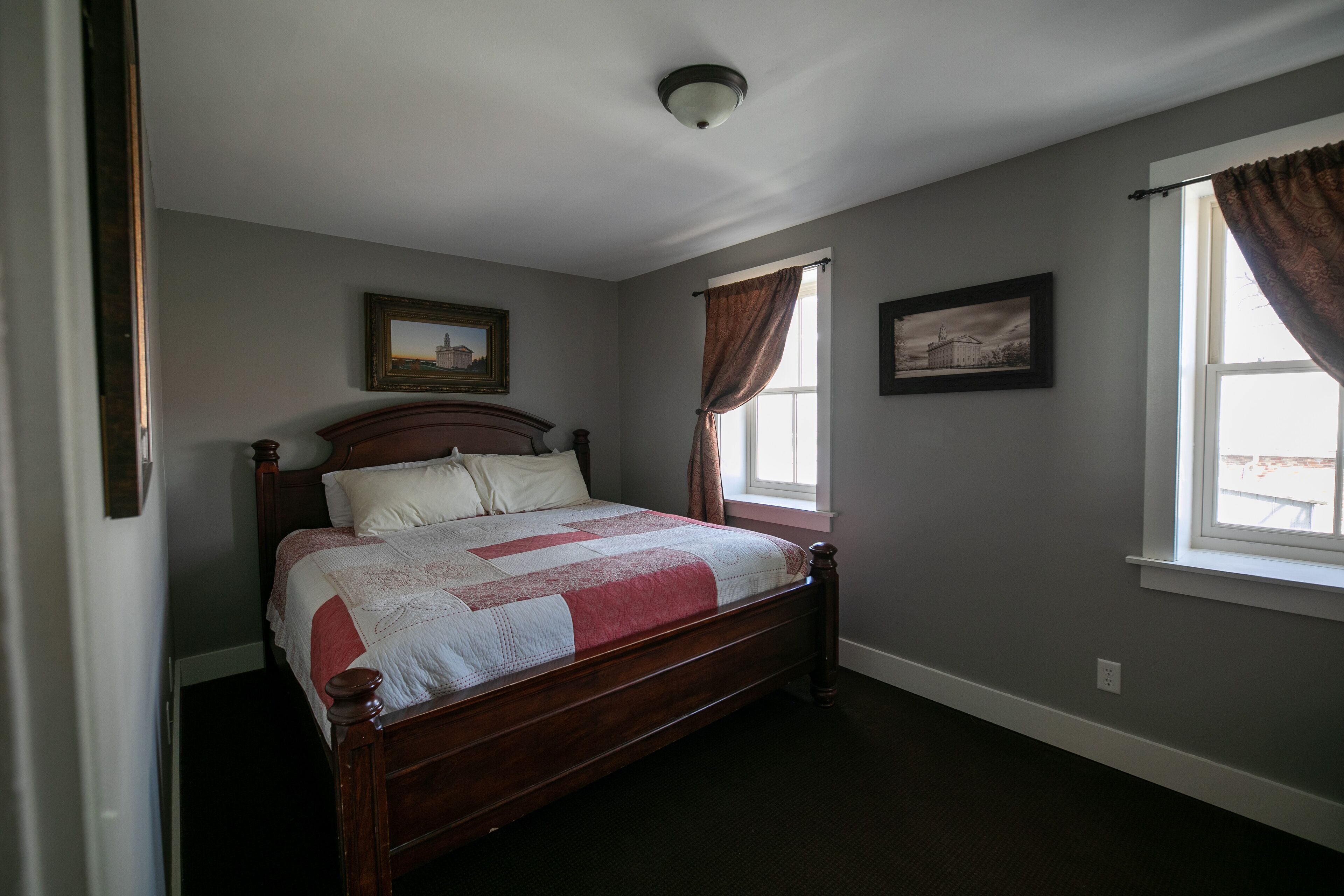Quarto com cama de madeira, duas quadros na parede, janela com cortinas e luminária no teto.