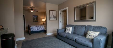 A living room with a grey sofa, a patterned bed, a ceiling fan, a framed picture, and a large mirror.