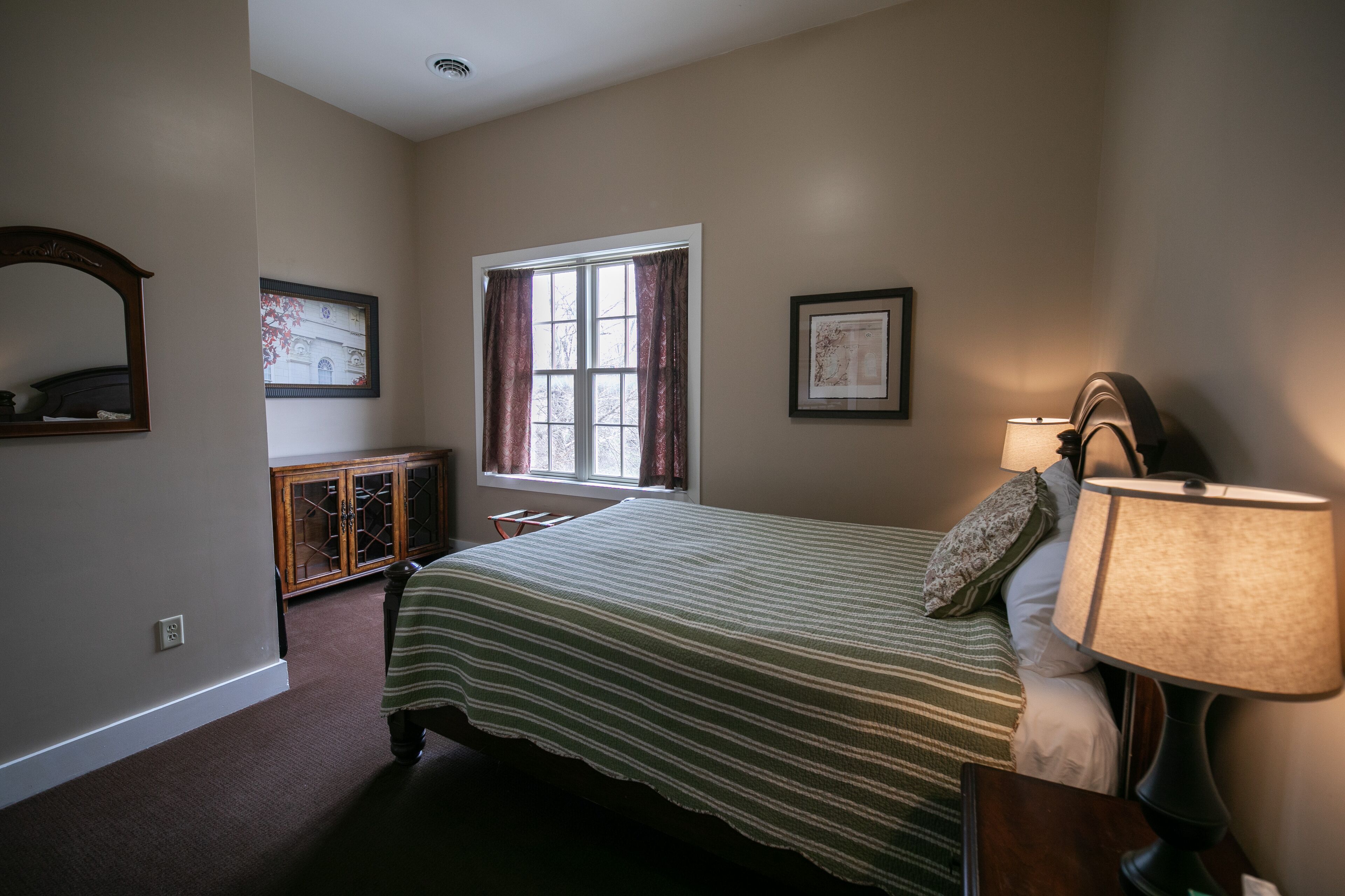 Une chambre à coucher avec un lit, une fenêtre avec des rideaux, une table de chevet avec une lampe, une commode et des tableaux encadrés accrochés au mur.