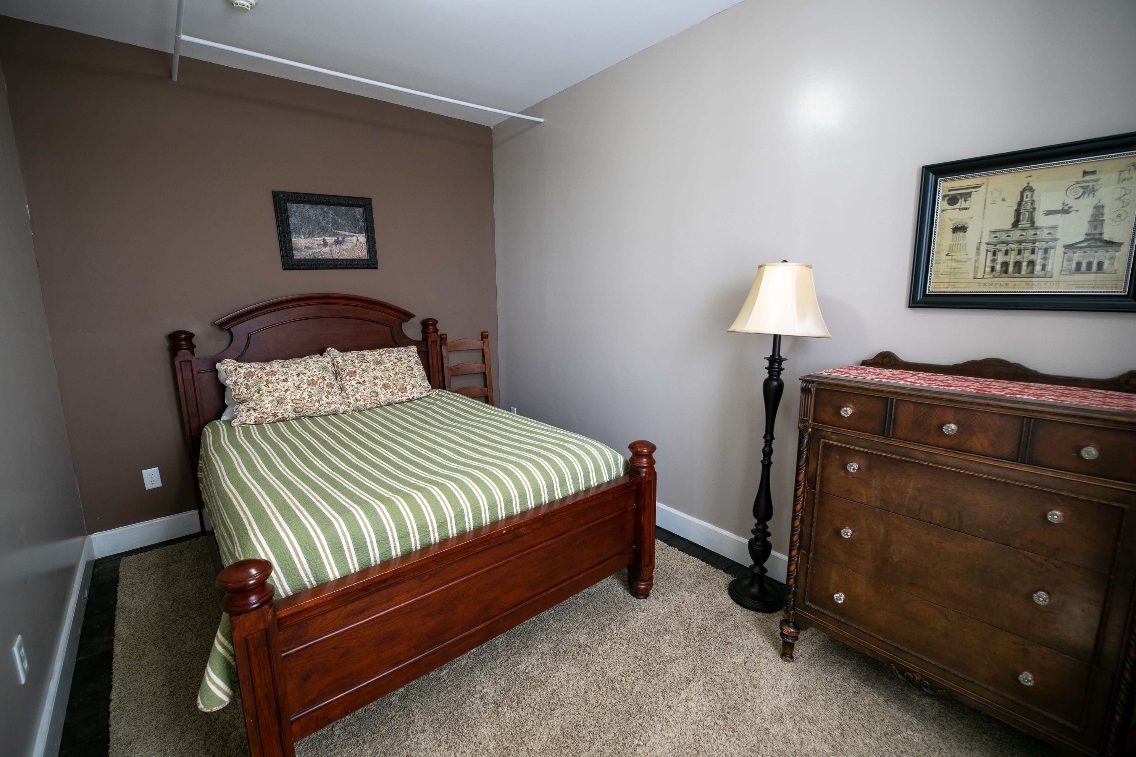 A bedroom with a wooden bed, a dresser, a lamp, and two framed pictures on the wall.