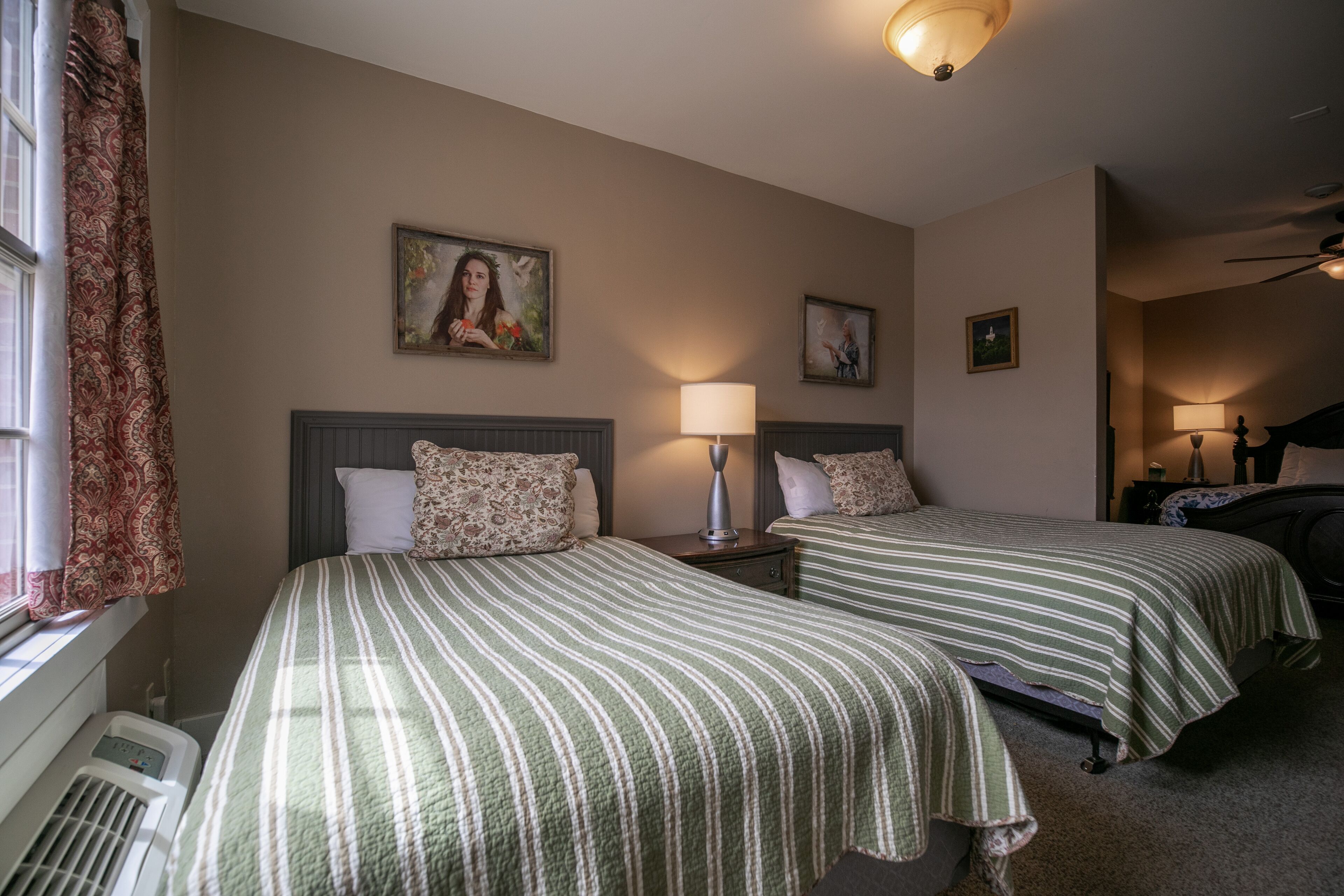 Une chambre d’hôtel avec deux lits, une fenêtre avec des rideaux, un radiateur et un ventilateur de plafond.