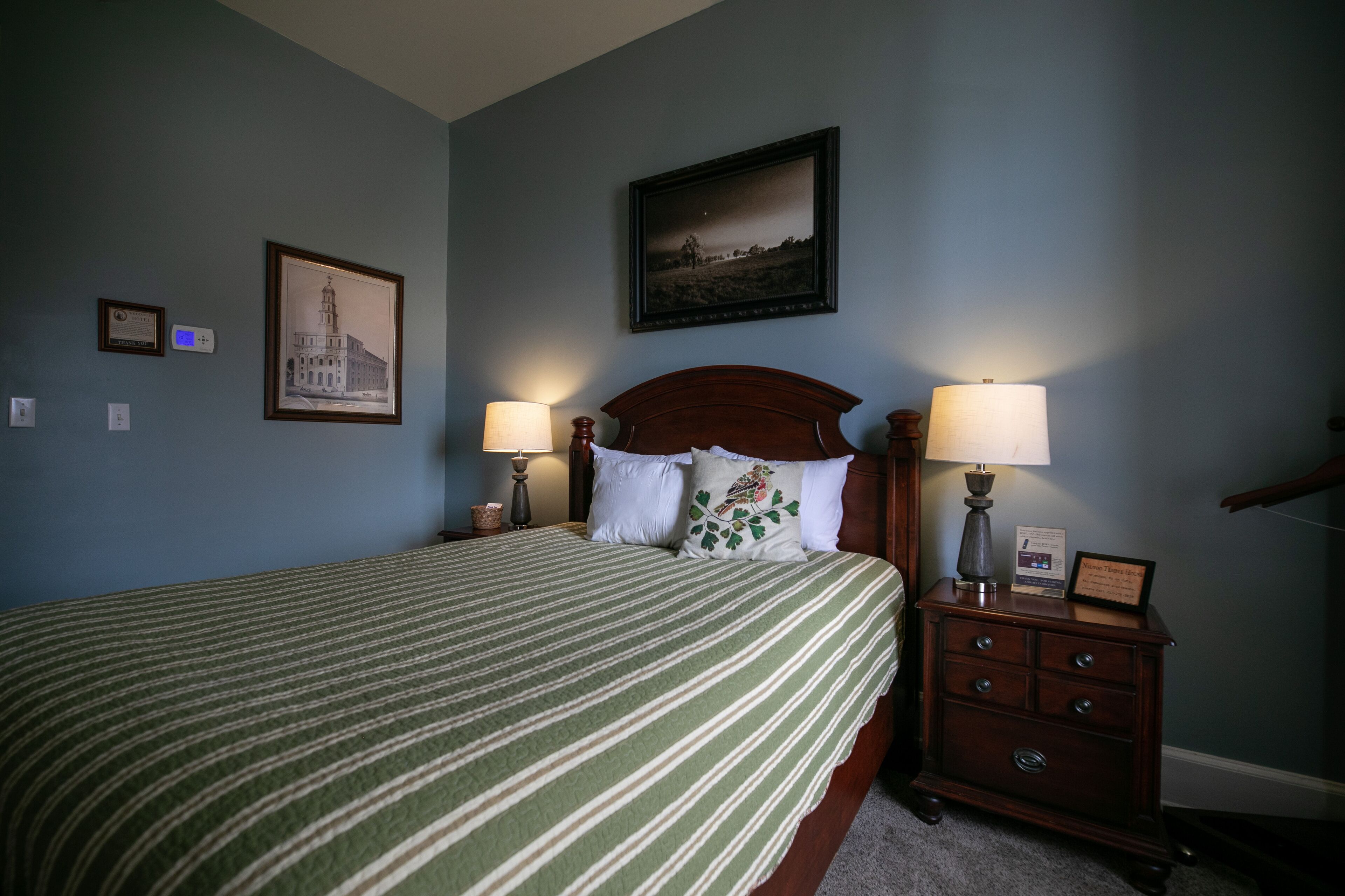 A neatly made bed with a striped bedspread, two bedside lamps, a framed picture above the bed, and a small dresser with a lamp on top.
