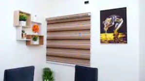 Une pièce dotée d’une grande fenêtre recouverte de stores, d’un fauteuil de couleur sombre, d’une petite table sur laquelle est posée une plante, et d’une œuvre d’art encadrée accrochée au mur.