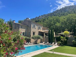 Une villa en pierre dotée d’une piscine, entourée d’une végétation luxuriante et adossée à un paysage montagneux.
